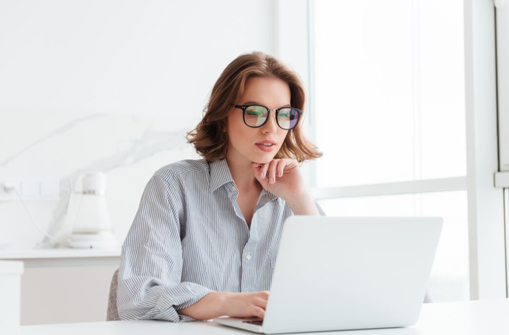 A focused woman in glasses working on her laptop in a bright office setting